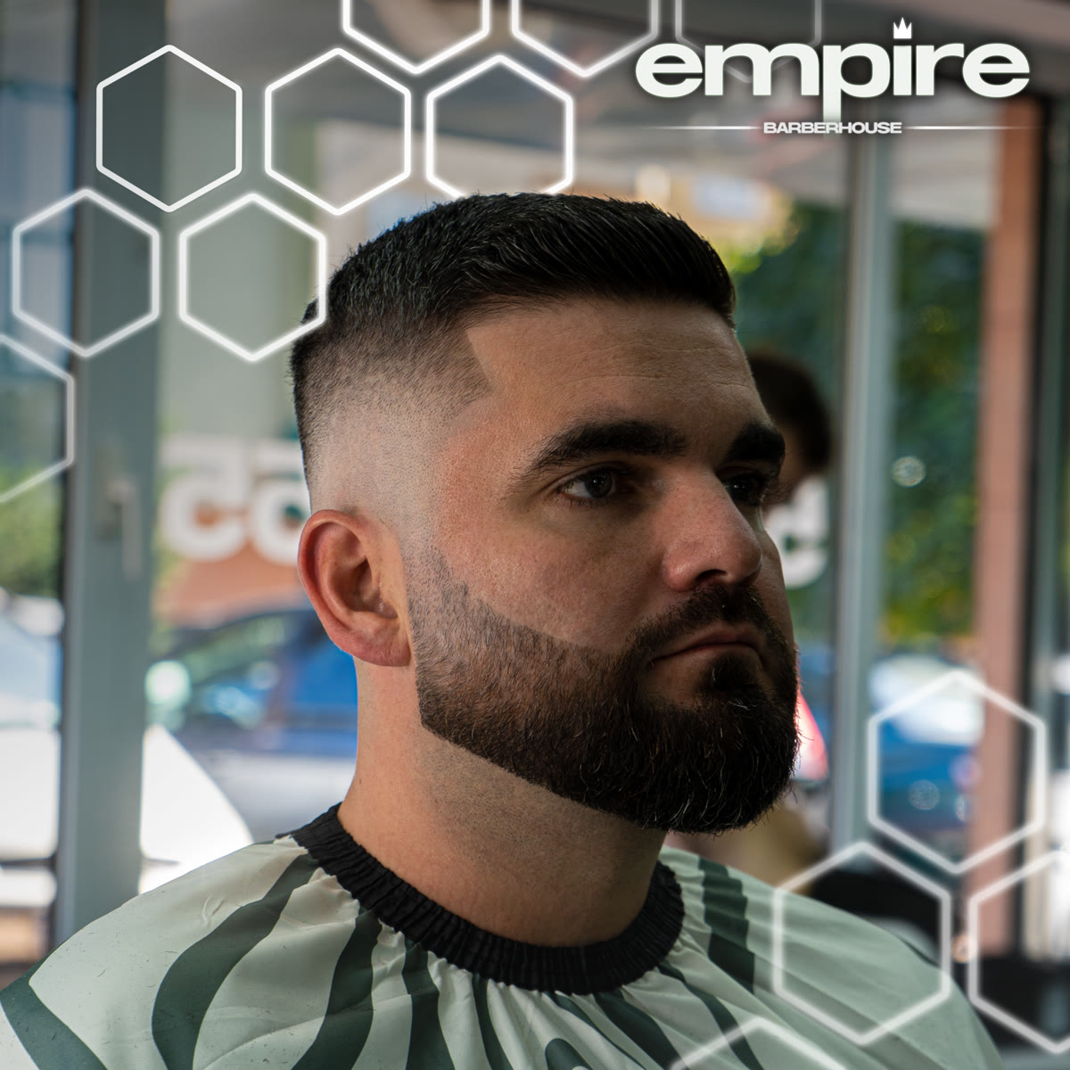 Wysoki skin fade z pełną, wyprofilowaną brodą — klient w pelerynie na tle salonu.